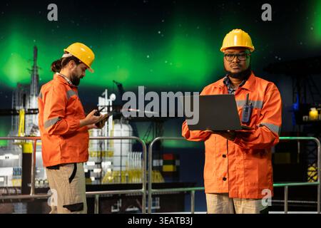 Ritratto di un esperto di barche di perforazione industriali che ispeziona l'integrità dell'attrezzatura utilizzando un computer portatile durante l'aurora boreale. Il lavoratore della piattaforma offshore esegue il controllo dei macchinari sotto l'aurora boreale verde Foto Stock