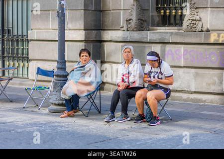 Tre donne sedute su sedie pieghevoli di fronte a un vecchio edificio, una che usa il suo telefono mentre aspetta pazientemente all'aperto. Foto Stock