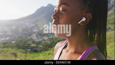 Donna in piedi che chiude gli occhi, testa inclinabile su una collina illuminata dal sole con trecce, parte superiore rosa, auricolari, spazio fotocopie Foto Stock