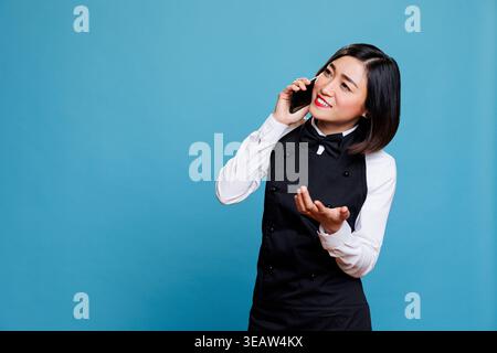 Sorridente giovane receptionist asiatica che parla al telefono cellulare, fornendo servizio ai clienti. Cameriera asiatica spensierata del ristorante vestita in uniforme mentre conversava sullo smartphone Foto Stock