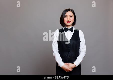Sorridente e attraente cameriera asiatica che indossa uniforme con cravatta di fiocco in piedi e guarda la macchina fotografica. Receptionist professionista del ristorante in posa per un ritratto in studio su sfondo grigio Foto Stock