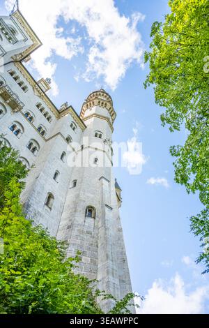 I visitatori si trovano vicino alla base del castello di Neuschwanstein, guardando le sue torri e le sue mura. Alberi verdi circondano il castello contro un cielo blu con nuvole. Foto Stock