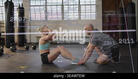 Tirocinante che indossa un reggiseno color acqua, allenatore che tiene i piedi in palestra con i kettlebell sovrapposti alla sala concerti Foto Stock