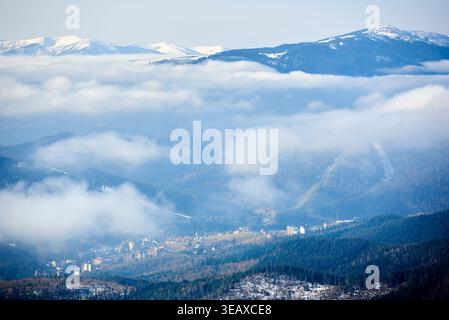 Pittoresca cittadina a valle annidata tra colline ondulate e fitte foreste. Valle inondata dalla luce soffusa del mattino, con nuvole che si spostano attraverso la scena. Le montagne innevate si innalzano sotto il cielo azzurro. Foto Stock