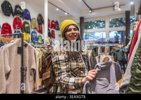 Giovane donna sorridente in un negozio alla moda di san francisco, che tiene in mano un capo d'epoca mentre si gode di una moda sostenibile, sfogliando un esclusivo prodotto di seconda mano Foto Stock