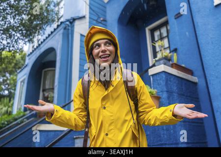 Donna che indossa un impermeabile giallo e un berretto sorridente mentre è in piedi sotto la pioggia, le mani si aprono per catturare le gocce di pioggia, con una facciata dell'edificio blu vivace in S. Foto Stock