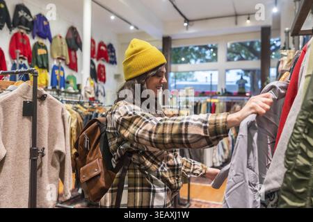 Donna che indossa un berretto giallo e una camicia a quadri che guarda attraverso una cremagliera di abbigliamento vintage in un vivace negozio thrift di san francisco, godendosi il selecti Foto Stock