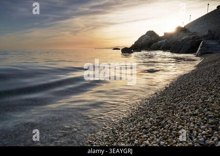 Tramonto sulla spiaggia di ciottoli di Varigotti, Liguria, Italia, con dolci onde mediterranee e una costa rocciosa inondata di luce dorata. Foto Stock