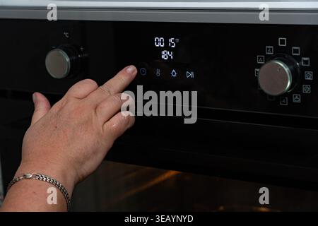 Vista ravvicinata di una mano che aziona il pannello di controllo digitale di un forno moderno, regolando la temperatura e le impostazioni di cottura per la precisione culinaria Foto Stock
