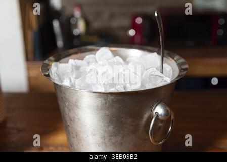 Un secchio metallico pieno di cubetti di ghiaccio si trova su un piano di legno, con uno sfondo della cucina. perfetta per bevande rinfrescanti o feste, aggiunge un tocco Foto Stock