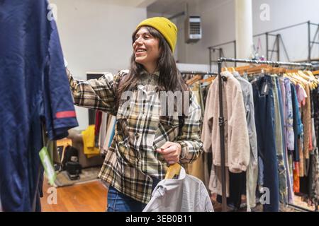Una giovane donna con una camicia a quadri e un berretto sorride mentre sfoglia i portapacchi in un negozio di articoli di abbigliamento di san francisco, scegliendo una mano di seconda mano unica e sostenibile Foto Stock