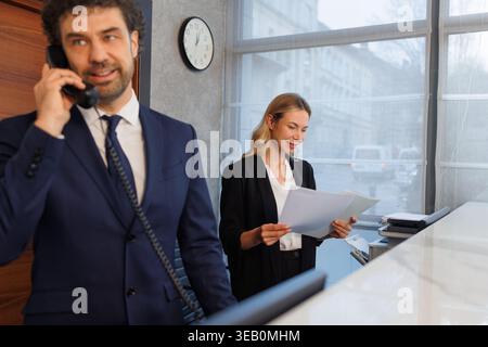 Uomo d'affari al banco della reception al telefono con assistente che legge i giornali dietro Foto Stock