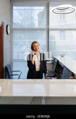 La receptionist dell'hotel è cordiale e parla al telefono dietro la reception nella hall moderna Foto Stock