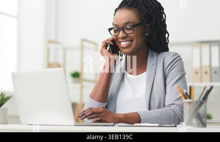 Una donna sorridente in una Gray Blazer partecipa a una chiamata telefonica mentre lavora su un notebook Foto Stock