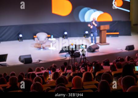 Relatore per conferenze sul palco che presenta al pubblico numeroso, telecamere professionali che registrano eventi dal vivo, moderna atmosfera da auditorium. Foto Stock