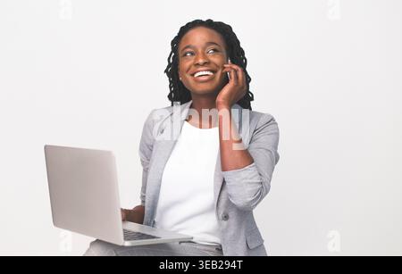 Donna sorridente che parla al telefono mentre utilizza il laptop in ambiente professionale Foto Stock