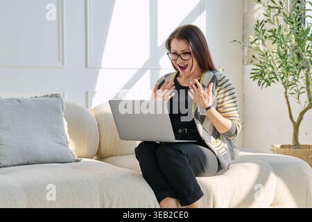 Donna matura sorridente con gli occhiali che videochiama con un computer portatile mentre si siede sul divano in un soggiorno luminoso. Concetto di lavoro remoto con terapia online o ascolto Foto Stock