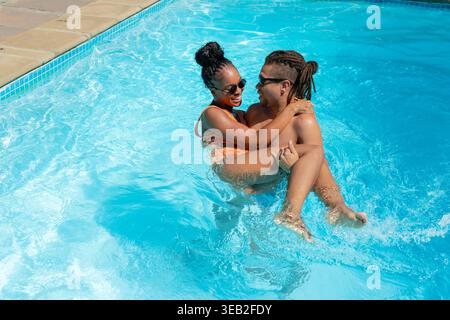Coppia afroamericana che si abbraccia mentre si tuffa nella piscina soleggiata con costumi da bagno e occhiali da sole, terrazza Foto Stock