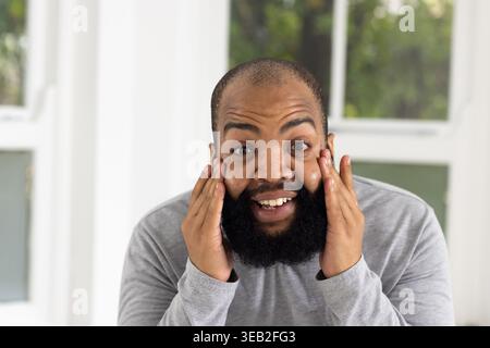Uomo africano che preme le palme alle guance, sorride accanto ai vetri della finestra, indossa una camicia grigio chiaro, barba intera Foto Stock