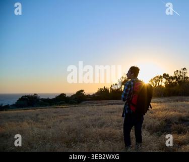 Uomo in backpacker che guarda il tramonto sull'oceano dalla collina Foto Stock