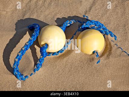 Due reti da pesca gialle intemprate aggrovigliate in una corda blu brillante riposano su una spiaggia di sabbia soffice sotto la calda luce del sole con ombre tenui e calma costiera. Foto Stock