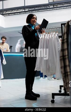 Dipendente del centro commerciale che gestisce l'inventario delle nuove collezioni di magliette utilizzando un tablet digitale. Giovane donna asiatica che controlla le scorte di abbigliamento in esposizione mentre lavora in un negozio di abbigliamento Foto Stock
