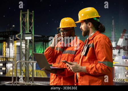 Membri dell'equipaggio della piattaforma offshore che utilizzano un laptop per controllare i sistemi sottomarini a tarda notte. I colleghi di lavoro di squadra monitorano il rilevamento delle anomalie utilizzando un notebook sul derrick di perforazione Foto Stock
