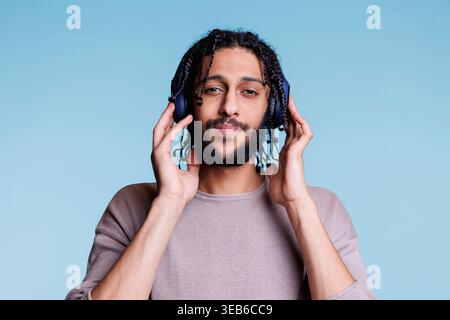 Un giovane arabo che tocca le cuffie sulla testa mentre ascolta musica, sorride e guarda la fotocamera. Persona che indossa cuffie wireless, goditi l'audio e i ritratti rilassanti Foto Stock