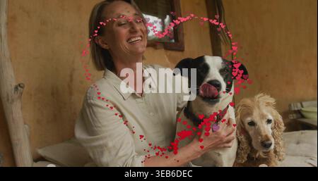 Donna sorridente in camicia beige che tiene due cani sul divano nel soggiorno, copricuore e collari Foto Stock