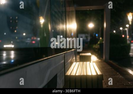 Scena urbana notturna con panchine in legno illuminate, marciapiedi bagnati che riflettono le luci della città, Foto Stock