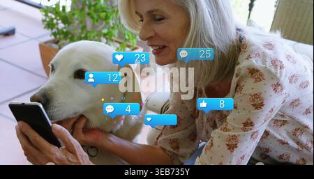 Donna anziana sorridente che indossa una camicetta rosa che tiene il telefono e coppetta il cane nella stanza del sole, bolle sociali Foto Stock