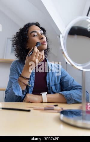 Persona non binaria che applica il trucco con la spazzola al tavolo da bagno utilizzando uno specchio rotondo con la parte superiore in denim Foto Stock