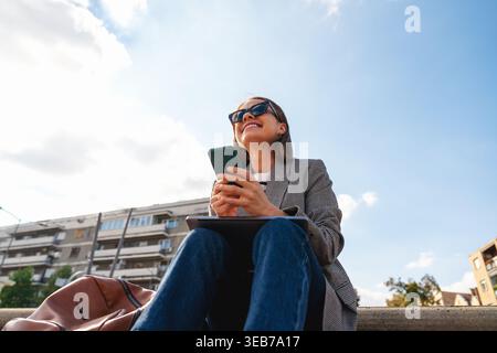 Vista ad angolo basso di una donna allegra che utilizza lo smartphone mentre si siede all'aperto sulla strada della città. Foto Stock