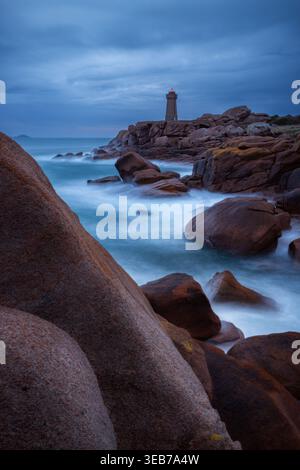 Veduta del faro di Ploumanaca'h che sorge su rocce di granito rosa con acqua di mare nebbiosa sotto un cielo al crepuscolo a Perros-Guirec, Bretagna, Francia. Foto Stock