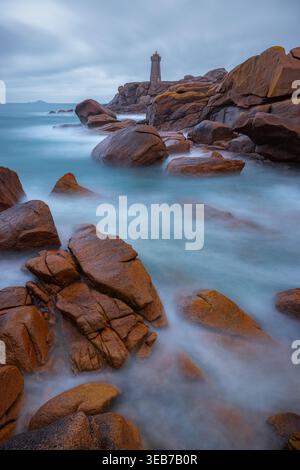 Vista del faro di Ploumanaca'h che si erge sulla Costa di granito rosa con lunghe onde di esposizione nebbiose che si infrangono contro grandi massi arancioni sotto una o Foto Stock