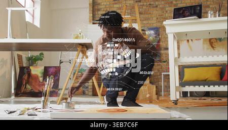 Artista maschile accovacciato che dipinge con pennello su tela da pavimento in studio, con cappuccio marrone, HUD visibile Foto Stock