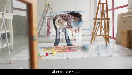 Artista donna che dipinge piegandosi su tela su un panno, tenendo tavolozza e spazzola, grembiule macchiato Foto Stock