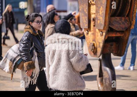 Batumi, georgia 21 aprile 2026: Una giovane donna con giacca di pelle e occhiali esamina un vecchio gancio arrugginito della gru mentre la compagna con cappotto di pelliccia cammina batumi p Foto Stock