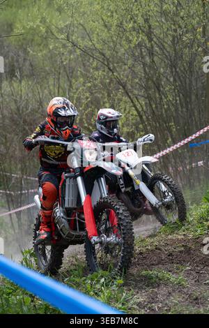 VLADIKAVKAZ, RUSSIA - 18 APRILE 2026: Due piloti di motocross in full gear che corrono su una pista sterrata attraverso una foresta durante una gara enduro. Foto Stock