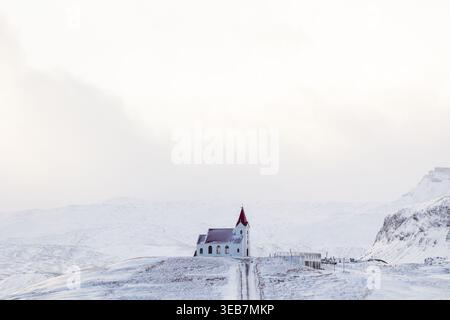 Vista di una piccola chiesa bianca con un tetto rosso che si erge su una collina innevata sullo sfondo di aspre montagne a Eyja- og Miklaholtshr, Islanda. Foto Stock