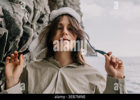 Donna cappello costa giovane donna che indossa un cappello bianco invernale in piedi vicino alla scogliera rocciosa del mare durante il viaggio autunnale Foto Stock
