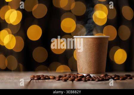 Bevanda calda al vapore in una tazza di carta bianca, circondata da chicchi di caffè tostati su un tavolo di legno, con calde luci bokeh Foto Stock