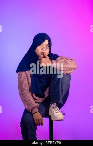 Una giovane donna siede premurosamente su uno sgabello. Indossa un hijab e un outfit casual. L'illuminazione viola e rosa crea una luce soffusa. La sua posa suggerisce calma Foto Stock