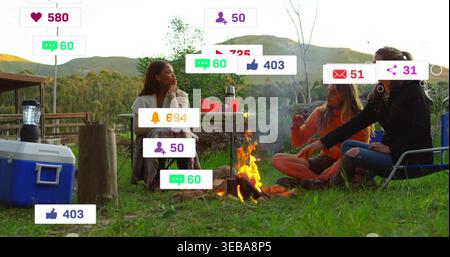 Raduna tre amici con foulard e la cima arancione intorno al fuoco del campeggio, al fresco e alle icone Foto Stock