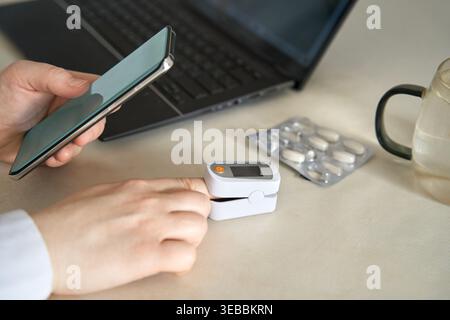 Pulsossimetro FingerTIP sul dito accanto a smartphone laptop e blister Pack che indica il controllo dello stato sulla superficie della scrivania. Foto Stock