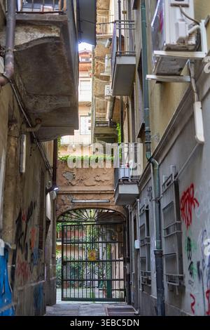 Un vicolo urbano stretto tra edifici residenziali invecchiati con pareti in cemento eroso, graffiti, balconi metallici e tubature a vista. La Foto Stock
