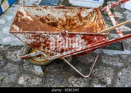 Carriola rossa e bianca usurata in piedi su ciottoli grigi, a simboleggiare l'età e il deterioramento Foto Stock