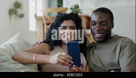 Coppia sorridente seduta su un divano leggero a casa, donna con la parte superiore beige che regge lo smartphone blu Foto Stock