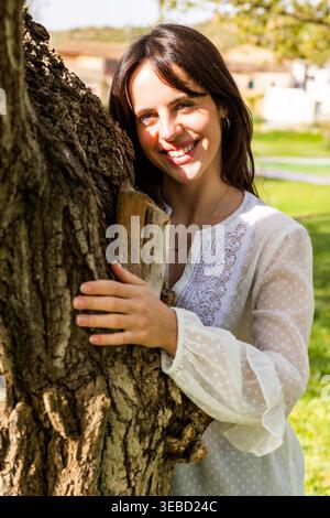 Giovane donna sorridente appoggiata al tronco dell'albero all'aperto Foto Stock