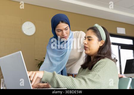 Diverse colleghe che si appoggiano su un laptop, rivedono il lavoro al tavolo dell'ufficio con l'orologio Foto Stock